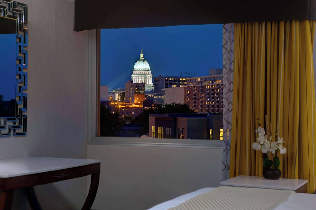 A room with a view of a cityscape, a window with curtains, a table, and a vase with flowers.
