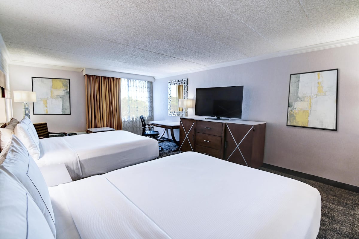 A hotel room with two beds, a desk, a television, and artwork on the walls.