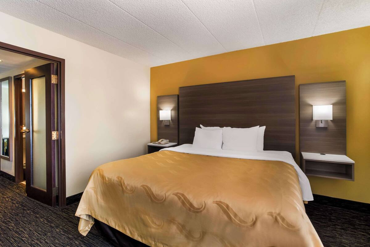 A hotel room with a large bed, two bedside tables with lamps, a door leading to another room, and a carpeted floor.