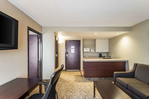 Una habitación de hotel con televisión de pantalla plana, una mesa de comedor, un sofá, una kitchenette y una alfombra estampada.