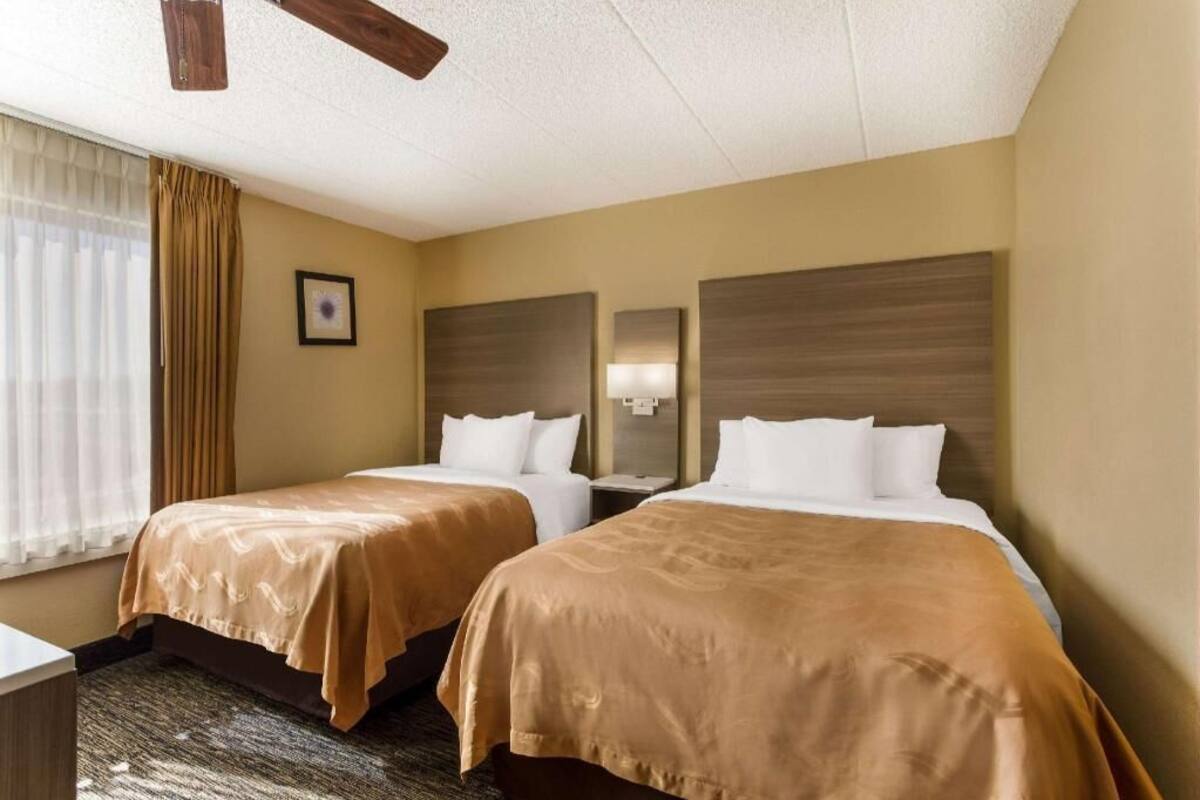 A hotel room with two beds, a ceiling fan, a window with curtains, and a framed picture on the wall.
