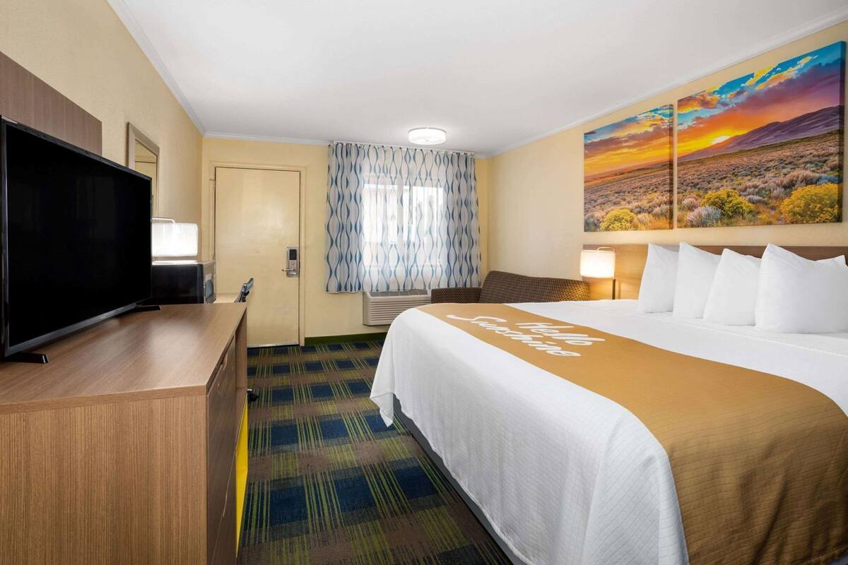 A hotel room with a bed, a television, a desk, a painting, and a patterned carpet.