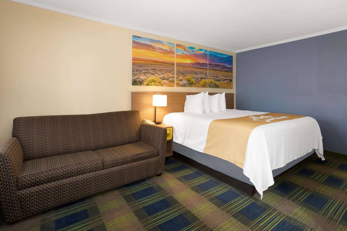 A hotel room with a bed, a sofa, a nightstand, a lamp, and a painting of a sunset landscape on the wall.