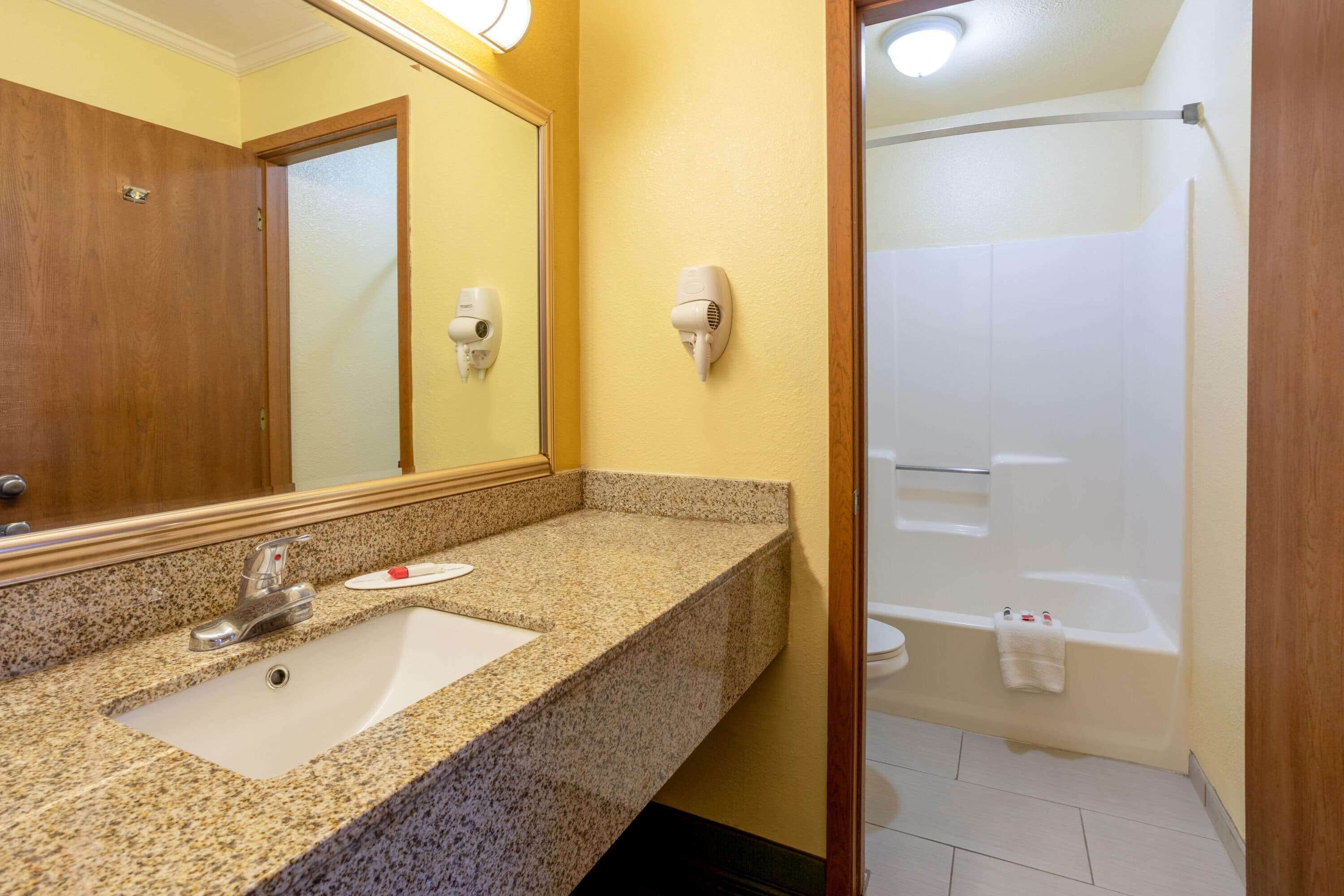 A bathroom with a granite countertop sink, a mirror, a hairdryer mounted on the wall, and a white bathtub.