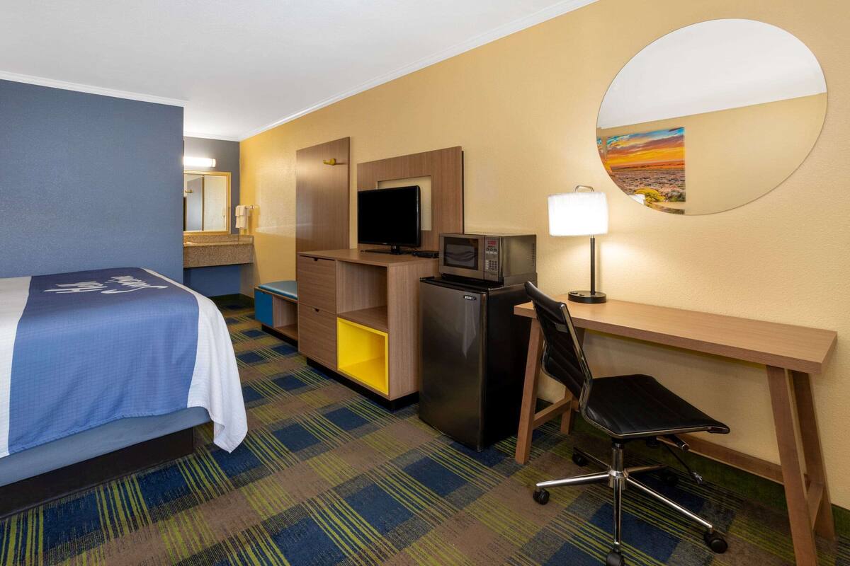 A hotel room with a bed, desk, chair, TV, refrigerator, and a mirror.
