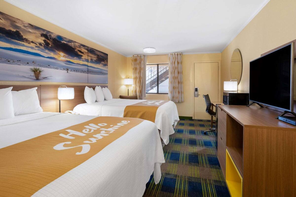 A hotel room with two beds, a TV, a desk, and a painting of a sunset.