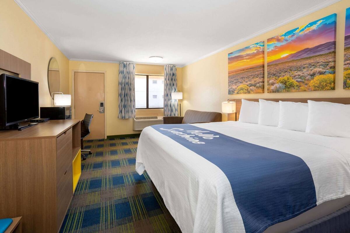 A hotel room with a large bed, a desk with a television, a window with curtains, and a painting on the wall.