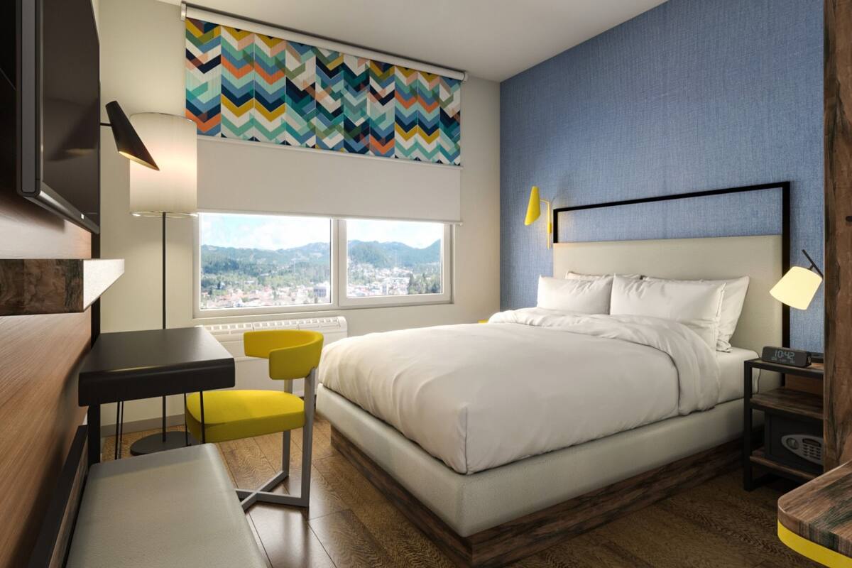 Ein Hotelzimmer mit Bett, Schreibtisch, Stuhl, Nachttisch, Lampe und Blick auf die Stadt durch ein Fenster.