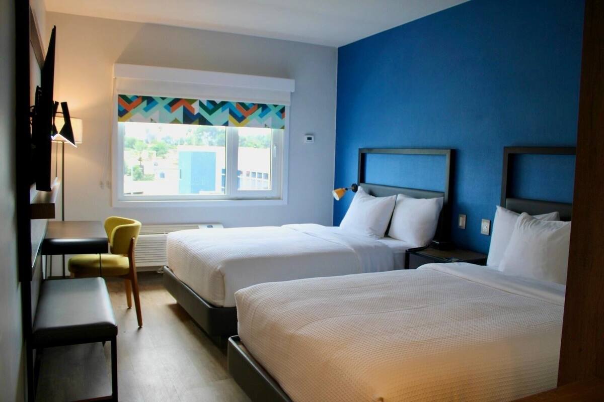 Ein Hotelzimmer mit zwei Betten, einem Schreibtisch, einem Stuhl und einem Fenster mit Blick auf Gebäude.