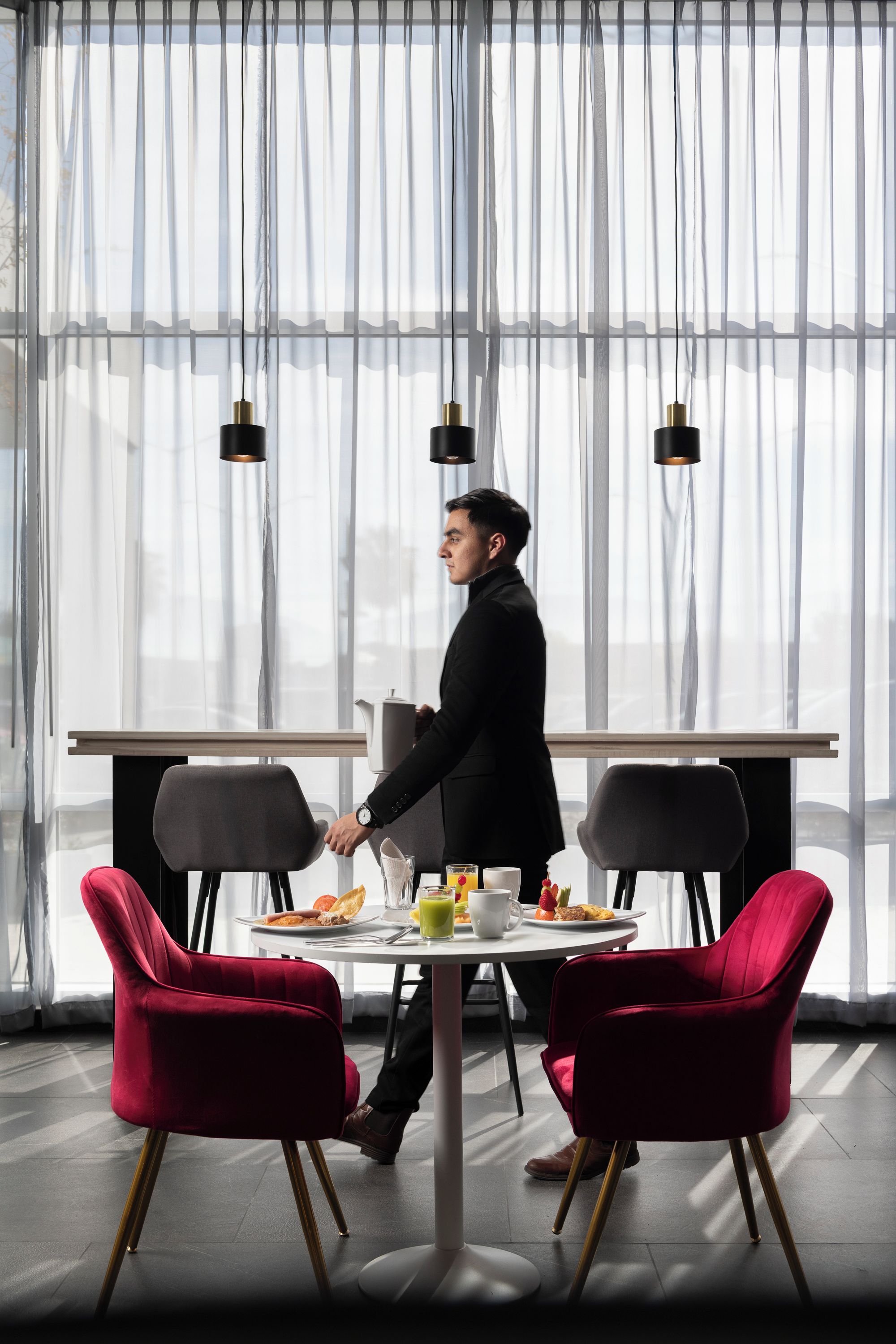 A modern dining area with a table set for two, featuring a white tablecloth, red chairs, and a coffee pot.
