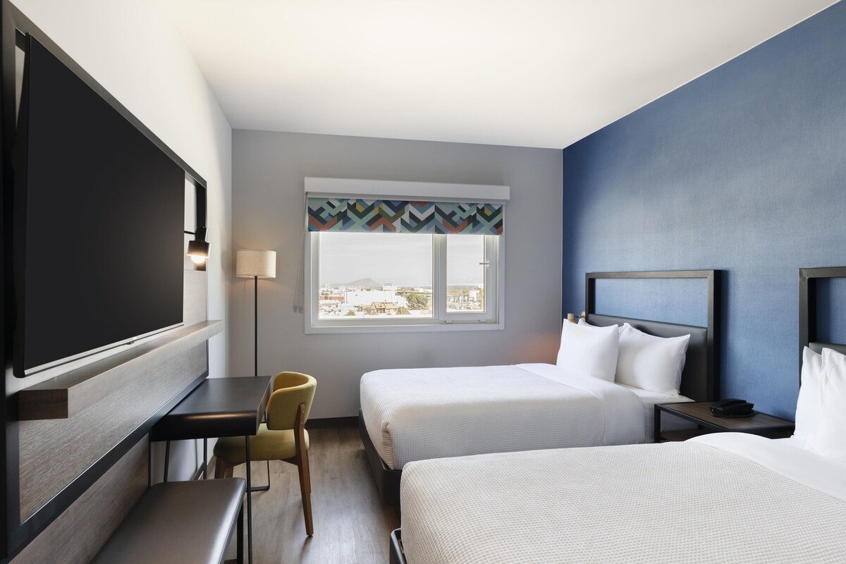 A hotel room with two beds, a desk, a chair, a TV, and a window with a view.