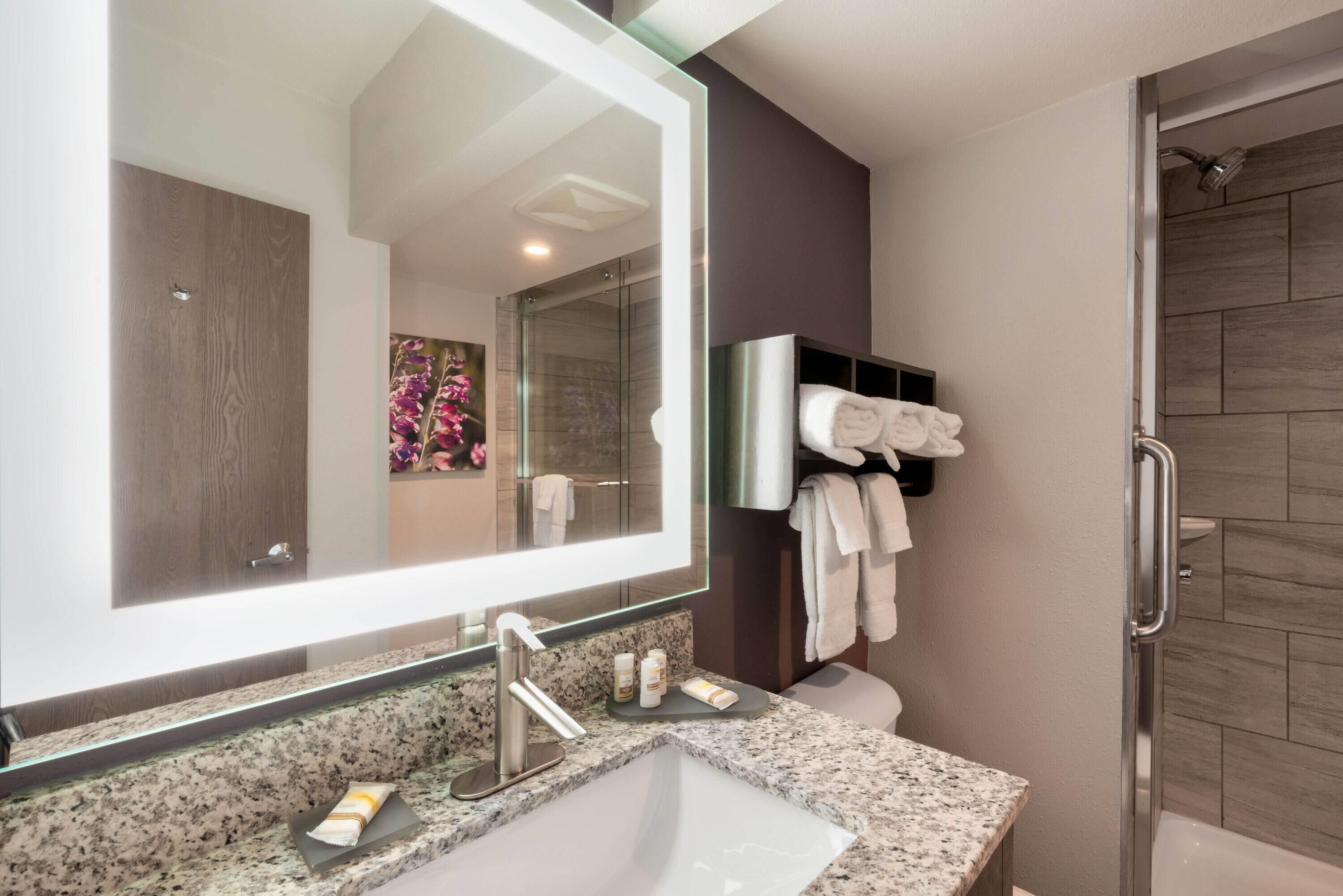 A modern bathroom with a granite countertop, a large mirror, a glass shower enclosure, and a towel rack with neatly folded towels.