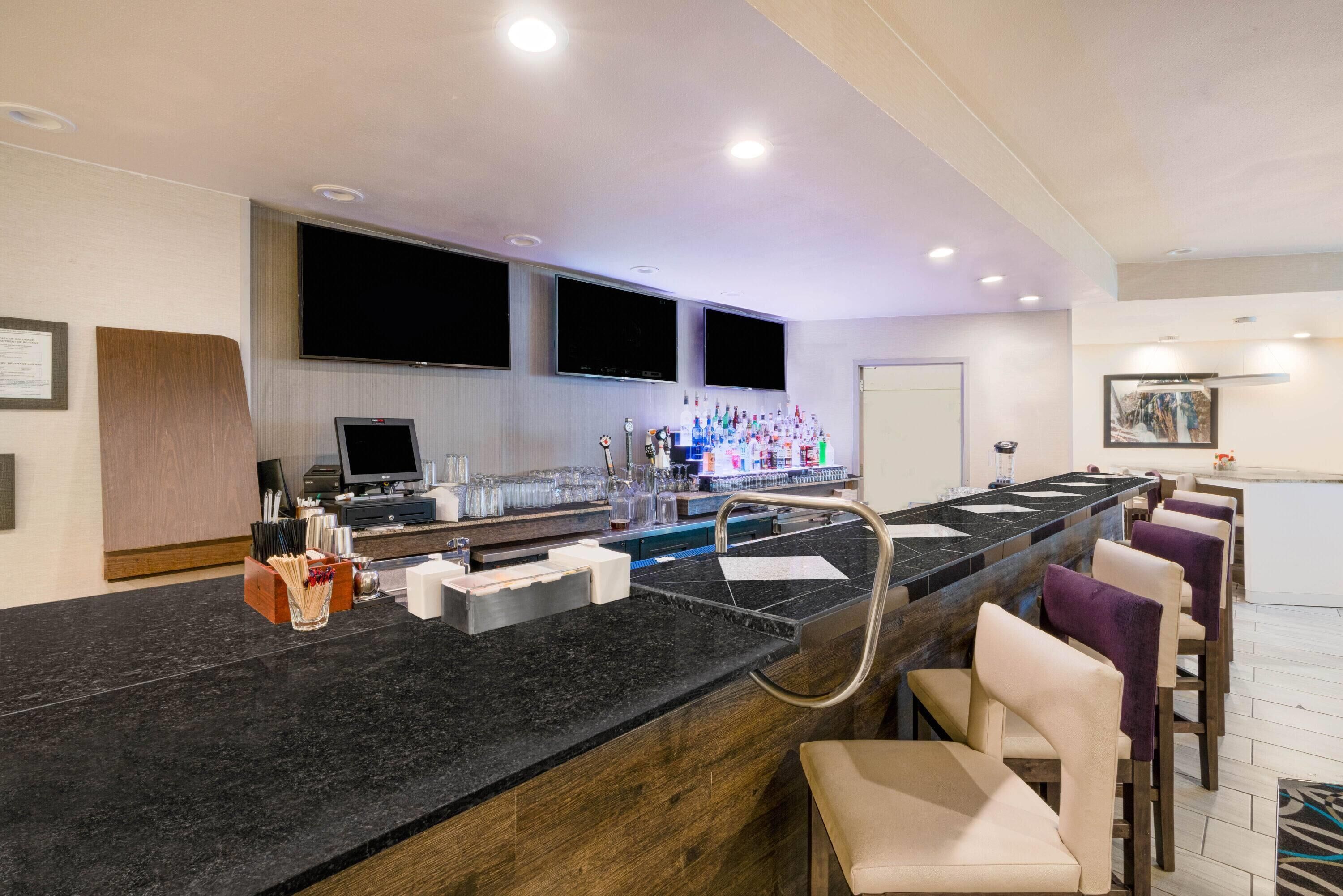 A modern bar area with a black countertop, multiple TVs, a bar stocked with bottles, and several cushioned bar stools.