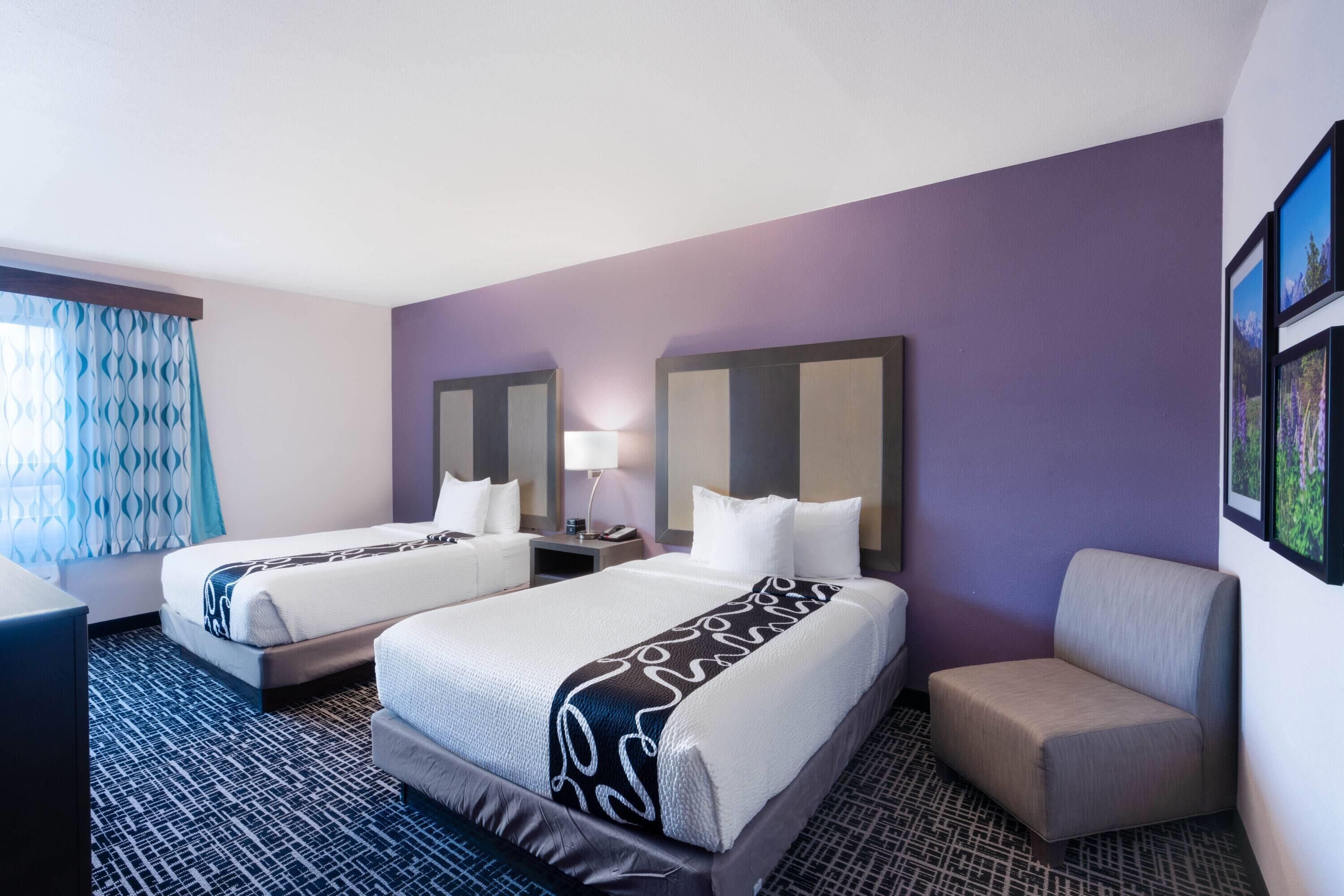 A hotel room with two beds, a desk, a chair, and a large window with patterned curtains.