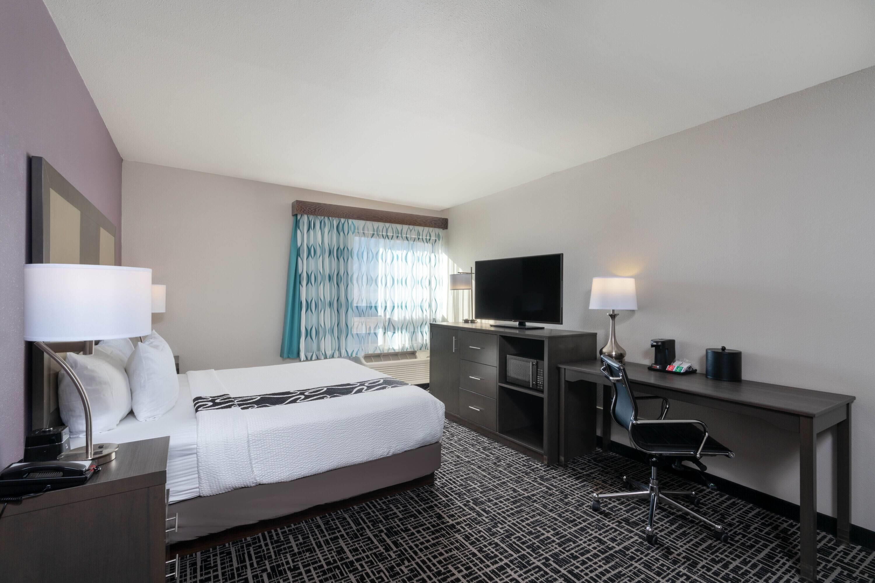 A hotel room with a bed, desk, TV, and a chair.