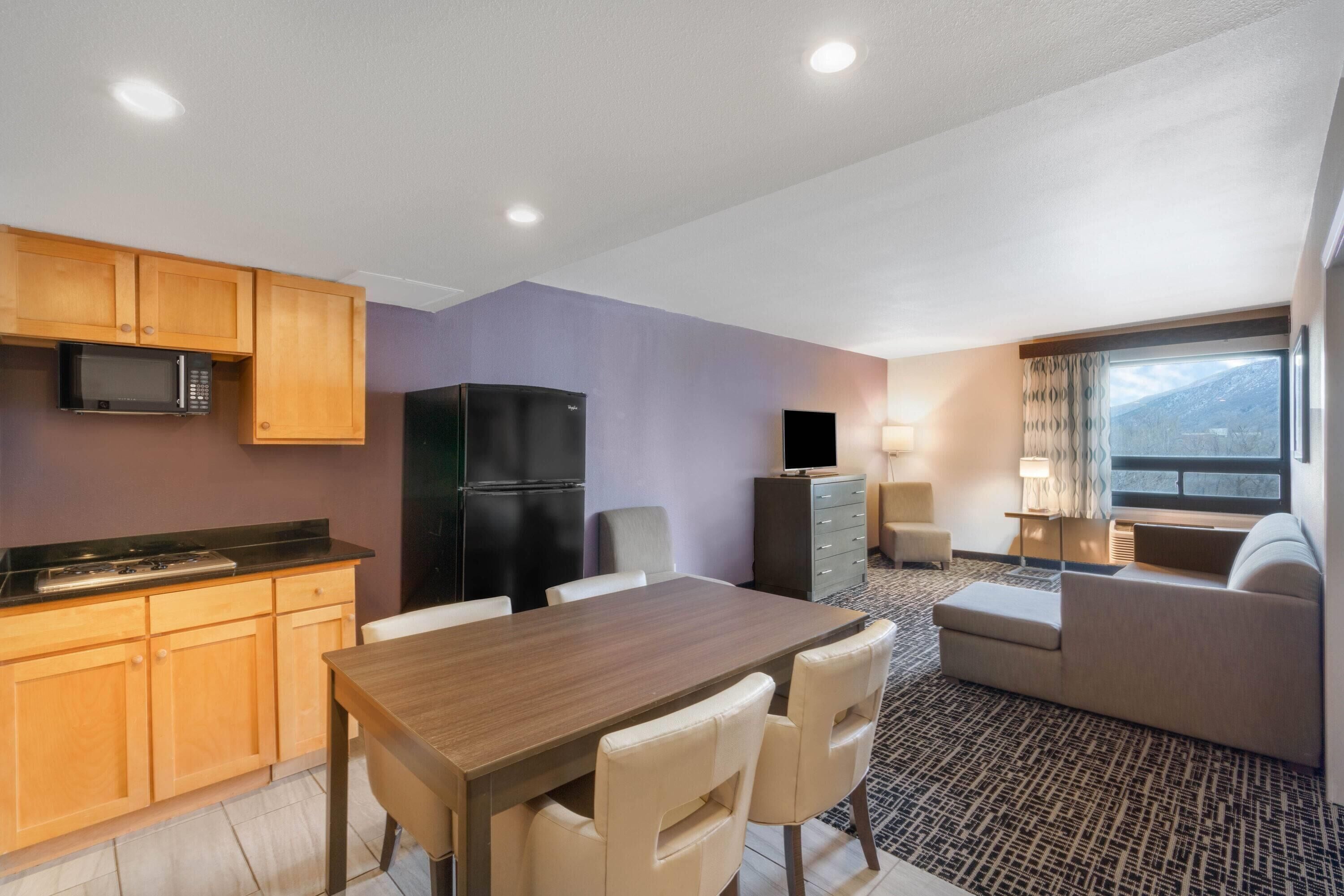 A modern hotel room with a dining area, kitchenette, and a sofa by a window.