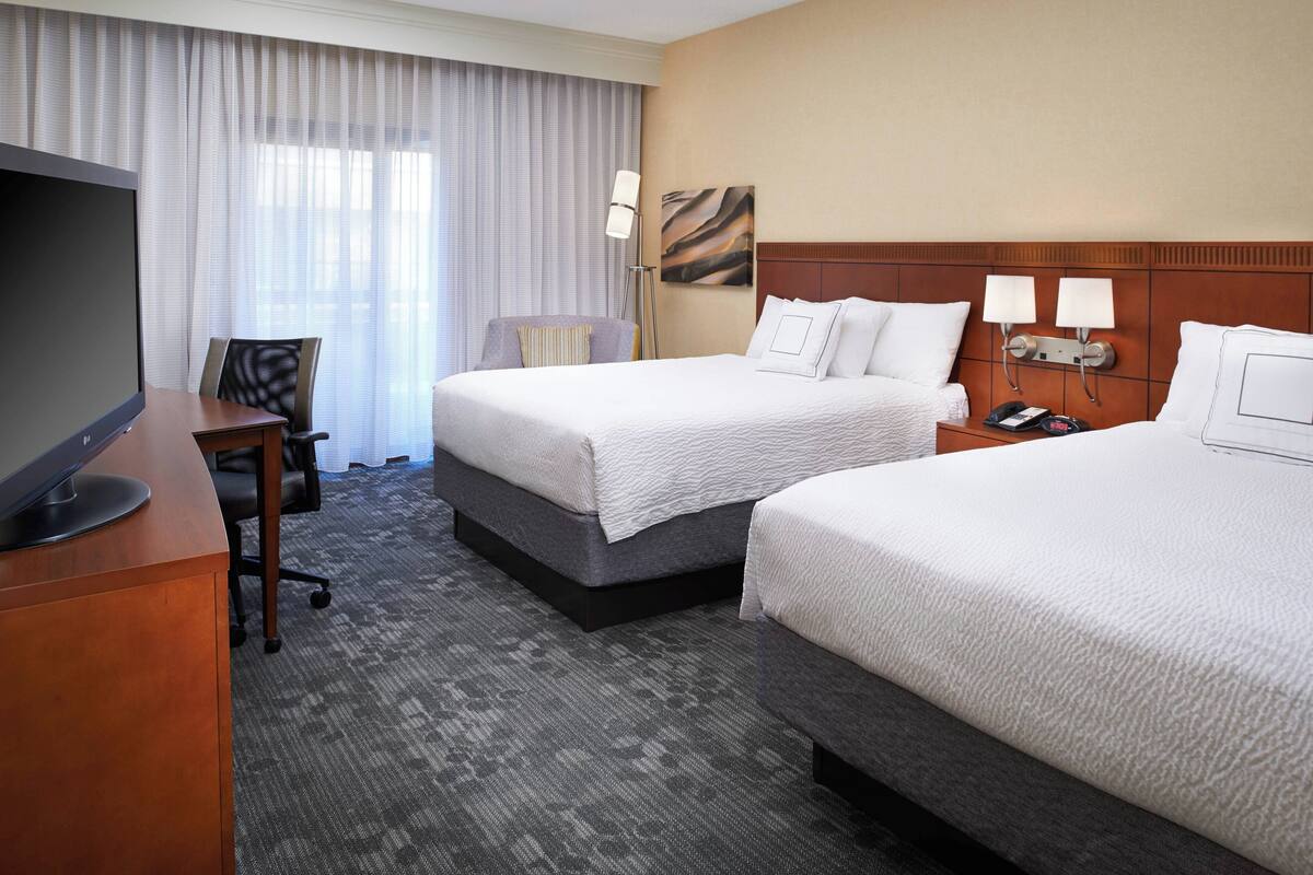 Ein Hotelzimmer mit zwei Betten, einem Schreibtisch mit Fernseher, einem Sessel und einem großen Fenster mit Vorhängen.