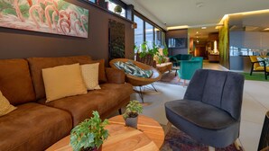 A modern living room with a brown sofa, teal armchairs, and a coffee table. There are potted plants and a painting of flamingos on the wall. - Novotel Nuernberg Messezentrum (Nuremberg)