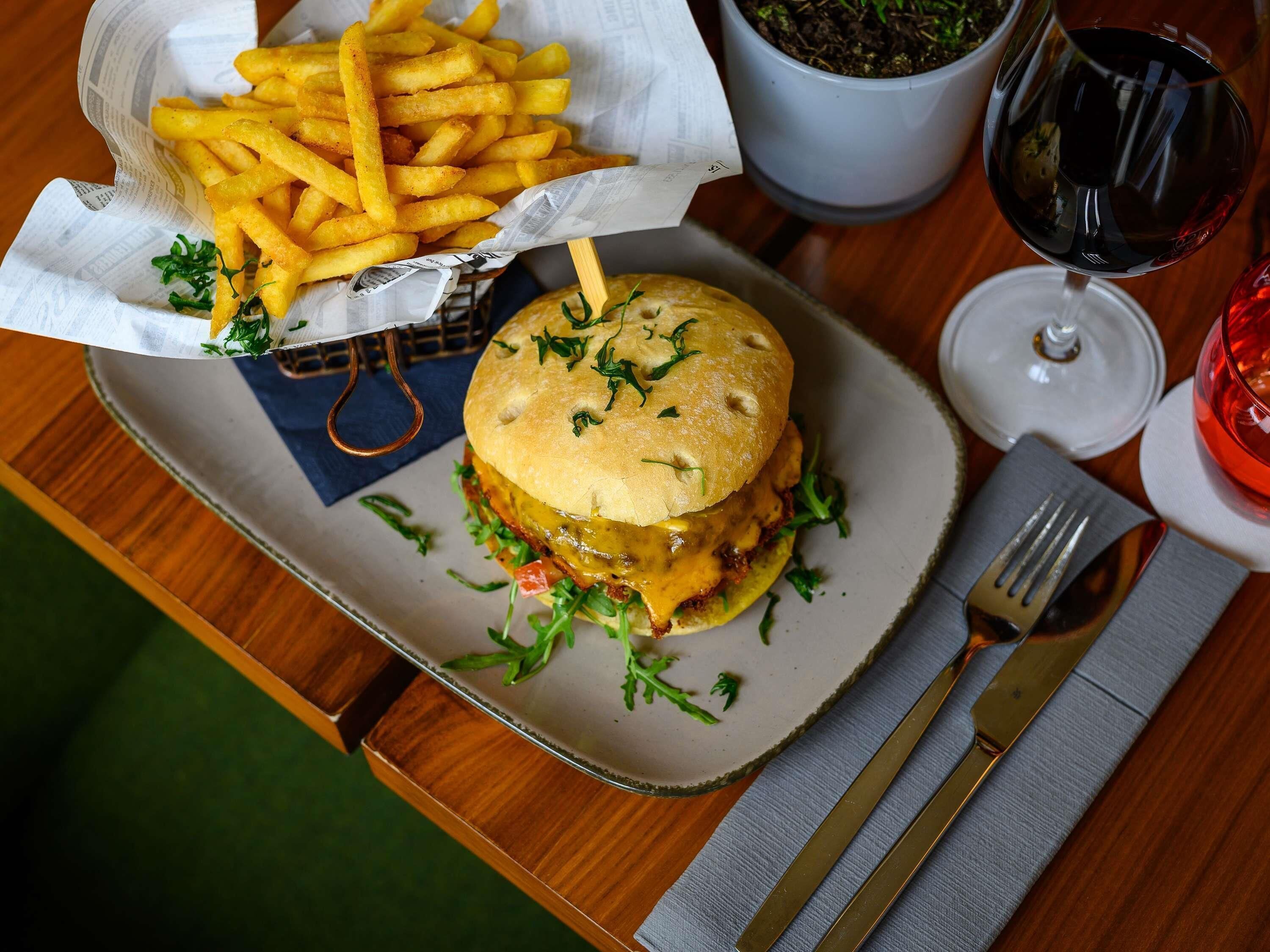 A meal with a burger and fries on a plate, a glass of red wine, and a napkin with cutlery on a wooden table.