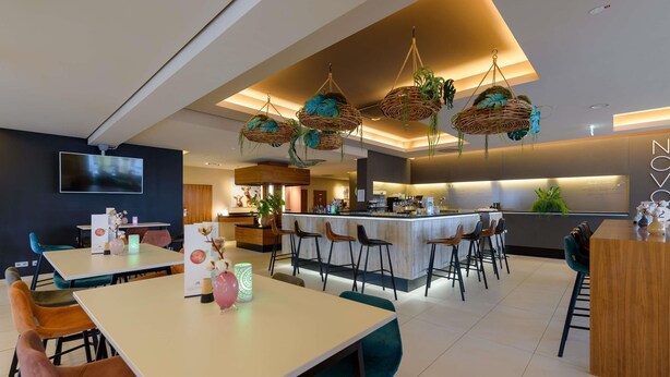 A modern restaurant with a bar area, multiple seating options, and a large TV screen. - Novotel Nuernberg Messezentrum (Nuremberg)
