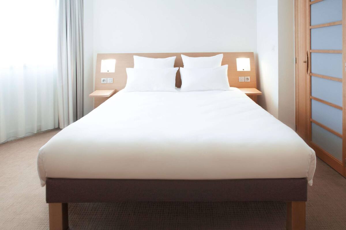 A hotel room with a large bed, wooden headboard, bedside lamps, and a view of the city through the window.