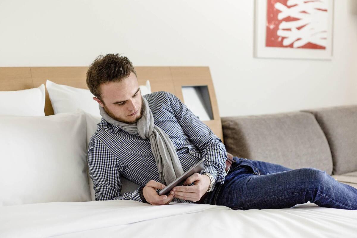A person lying on a bed with a tablet, wearing a checkered shirt and jeans, in a room with a wooden headboard and a framed picture on the wall.