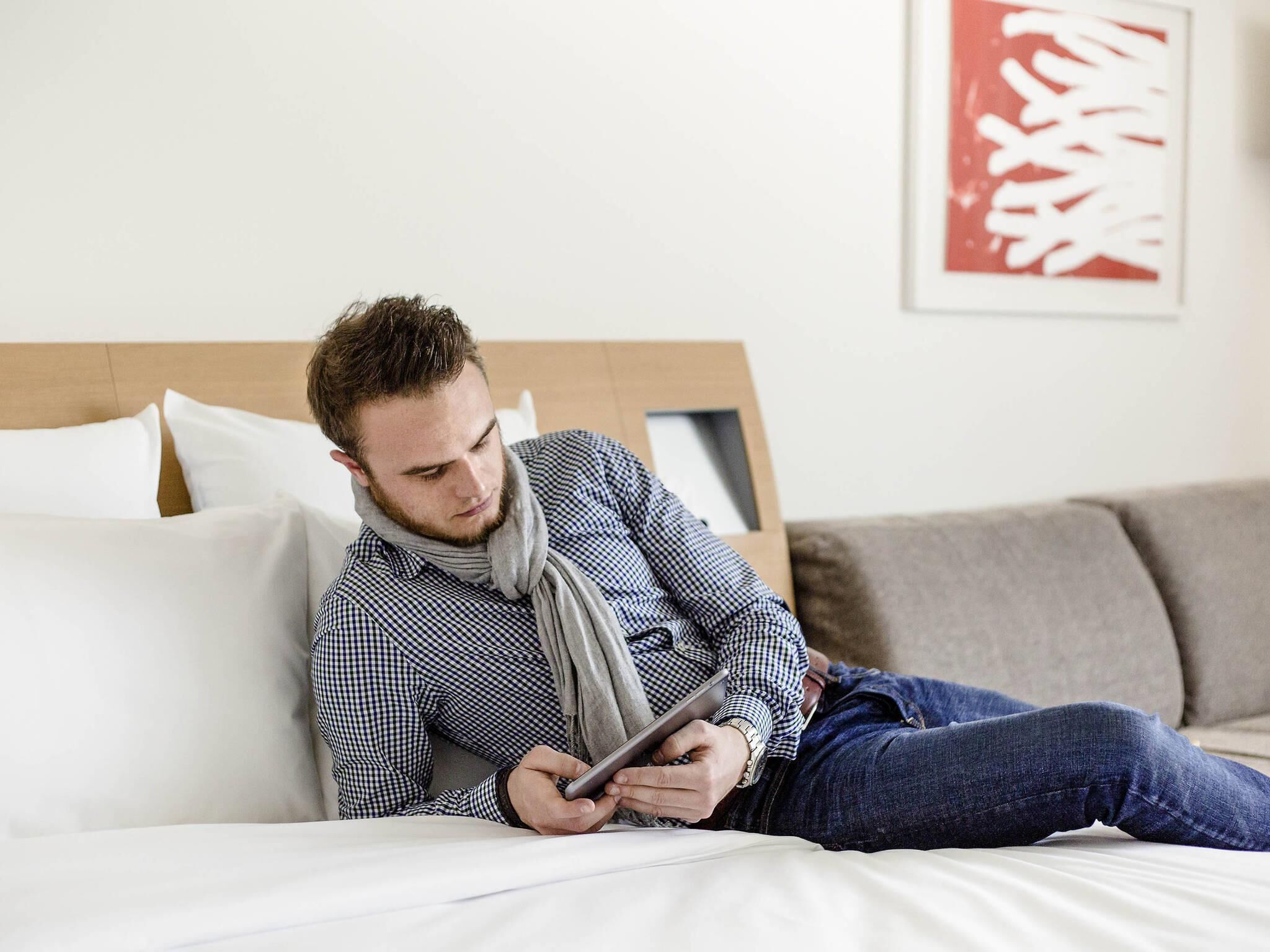 A person lying on a bed with a tablet, wearing a checkered shirt and jeans, in a room with a wooden headboard and a framed picture on the wall.