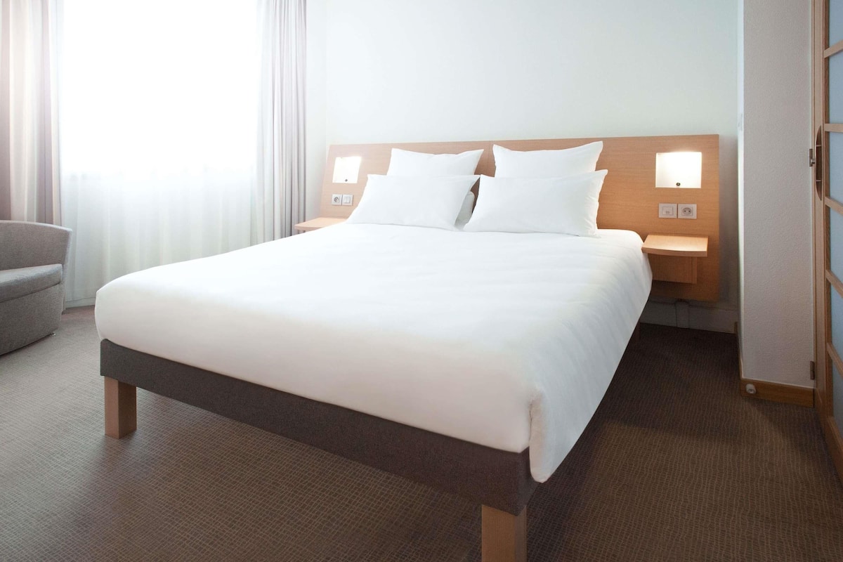 A hotel room with a large bed, two bedside tables, a chair, and a window with curtains.