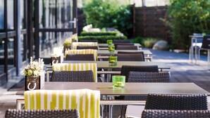 An outdoor dining area with tables and chairs, a glass display case with flowers, and a view of greenery. - Novotel Nuernberg Messezentrum (Nuremberg)