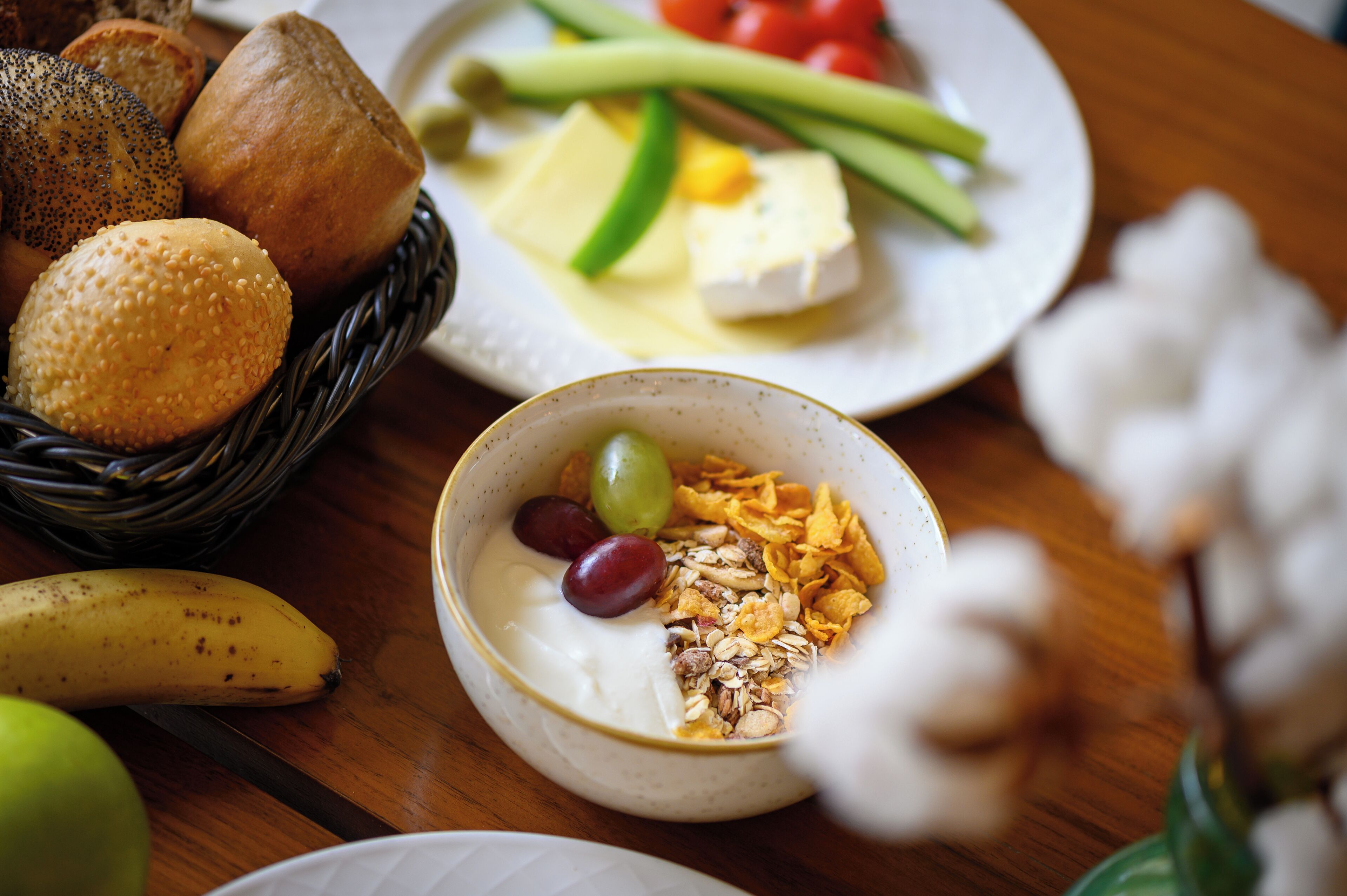 A table with a variety of food items including bread, cheese, vegetables, and a bowl of yogurt with granola and grapes.