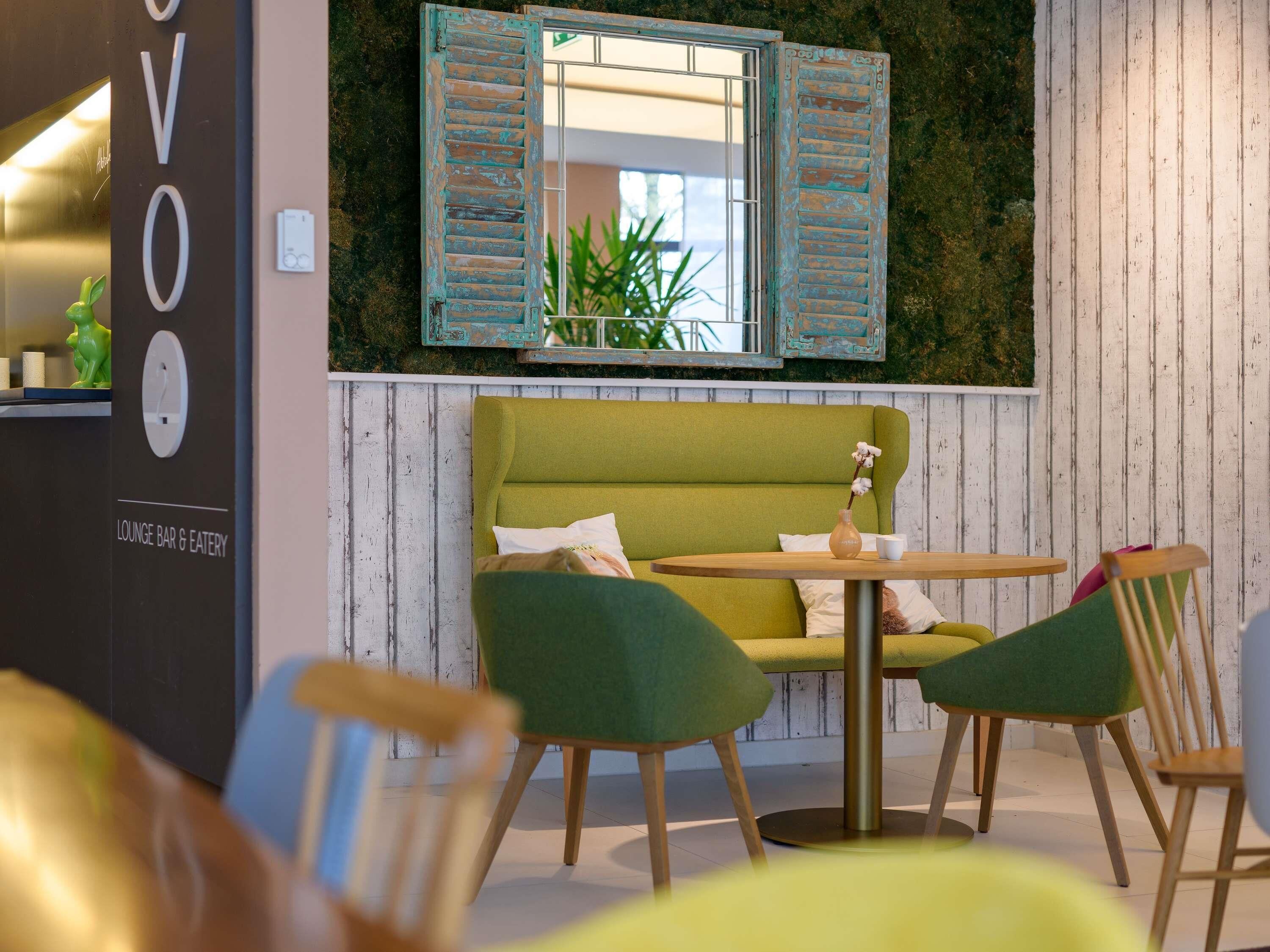 Un área de descanso con un sofá verde, sillas de madera, una mesita y un espejo con contraventanas.