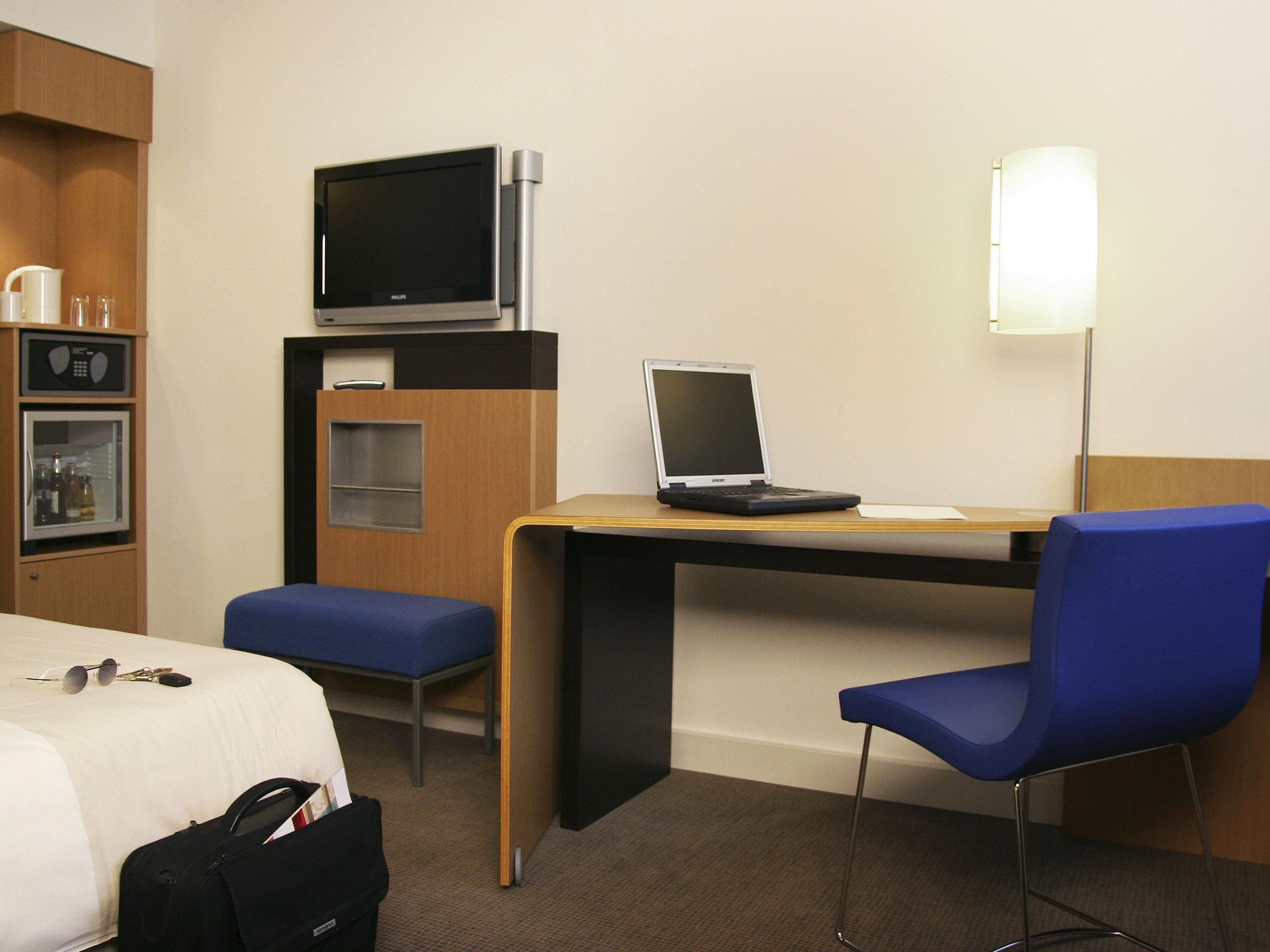 Habitación de hotel con cama, escritorio con una computadora portátil, silla, televisor, minibar y lámpara.