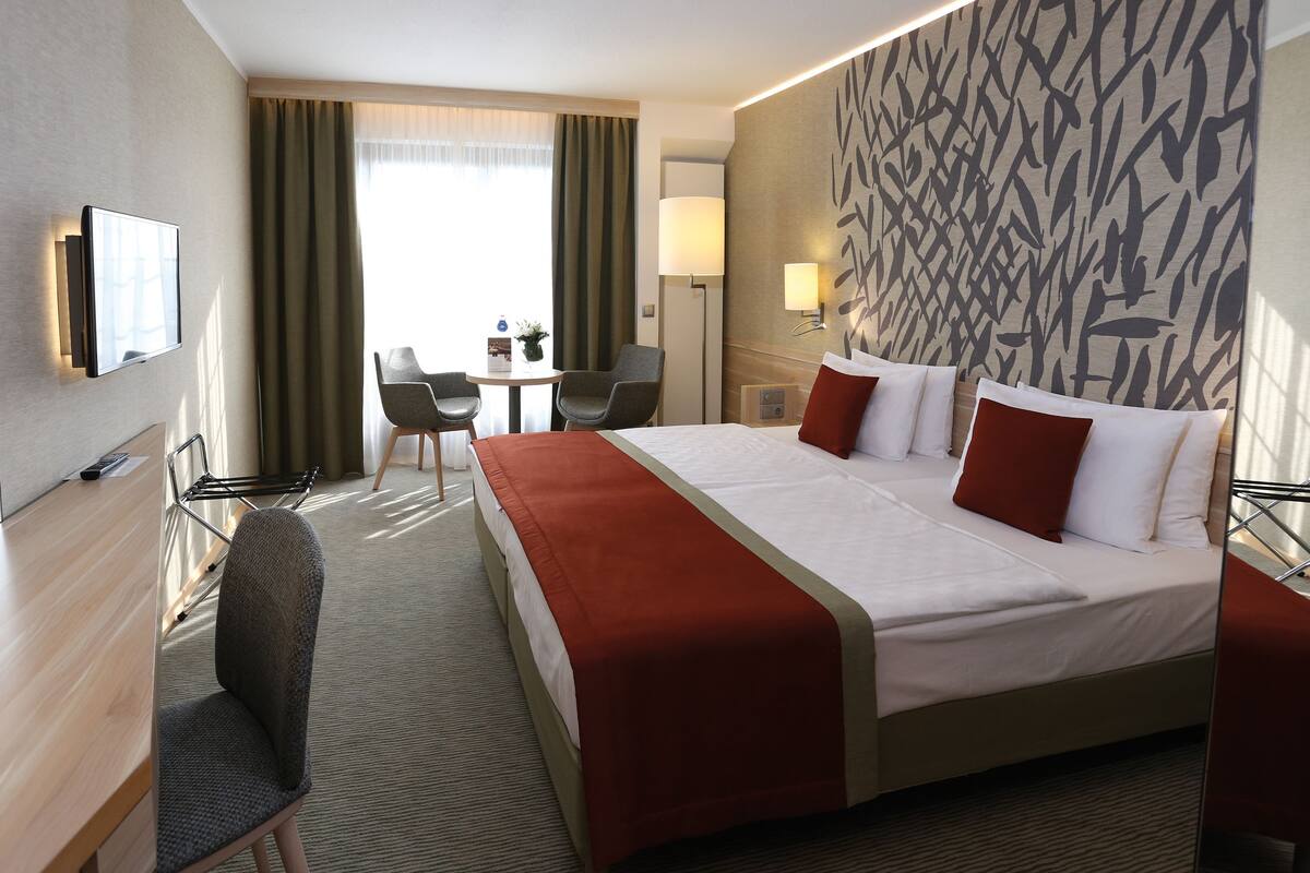 A hotel room with a large bed, a desk with a chair, a TV, and a view of the outdoors.