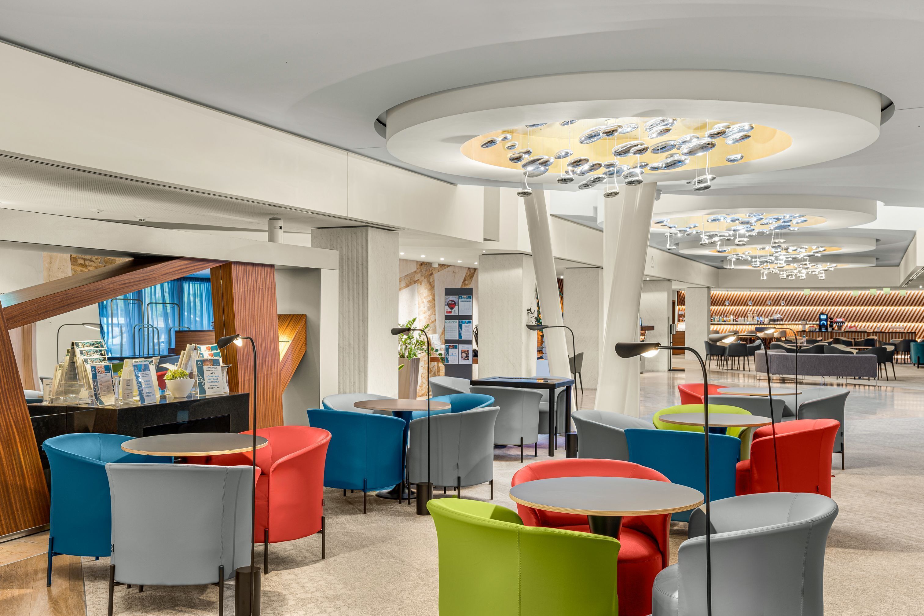 A modern interior with round tables and colorful chairs, a central light fixture, and a reception area with a counter and seating.