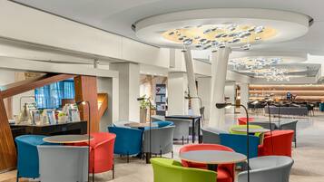 A modern interior with round tables and colorful chairs, a central light fixture, and a reception area with a counter and seating.