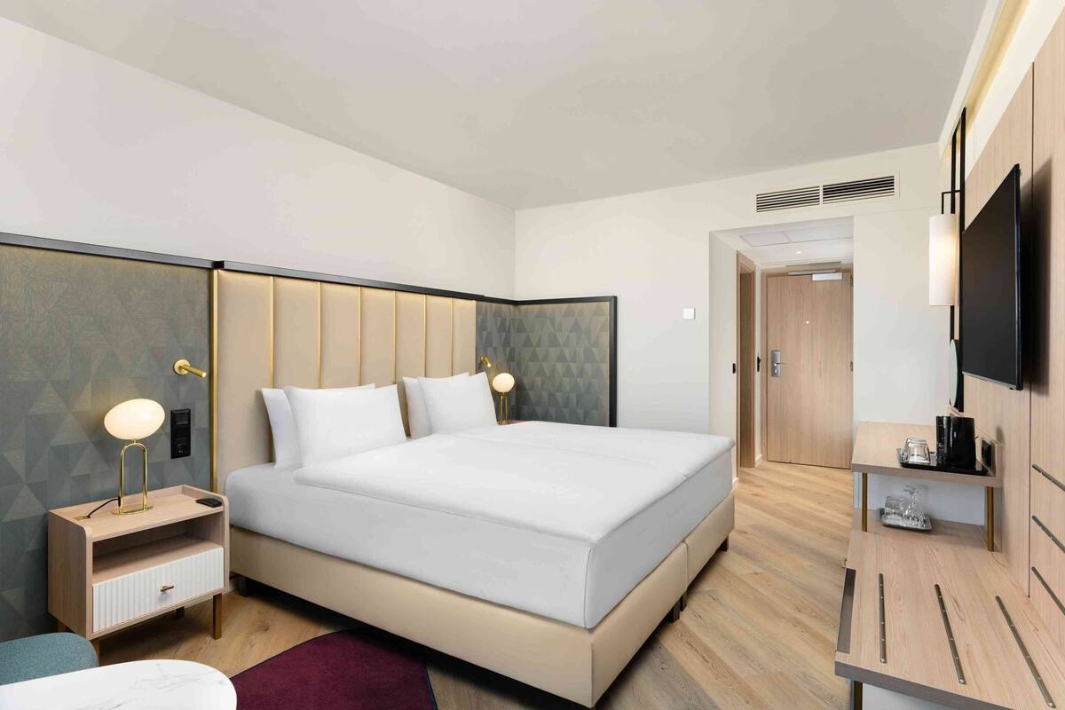 A modern hotel room with a large bed, bedside tables with lamps, a TV, and a small desk with a kettle.