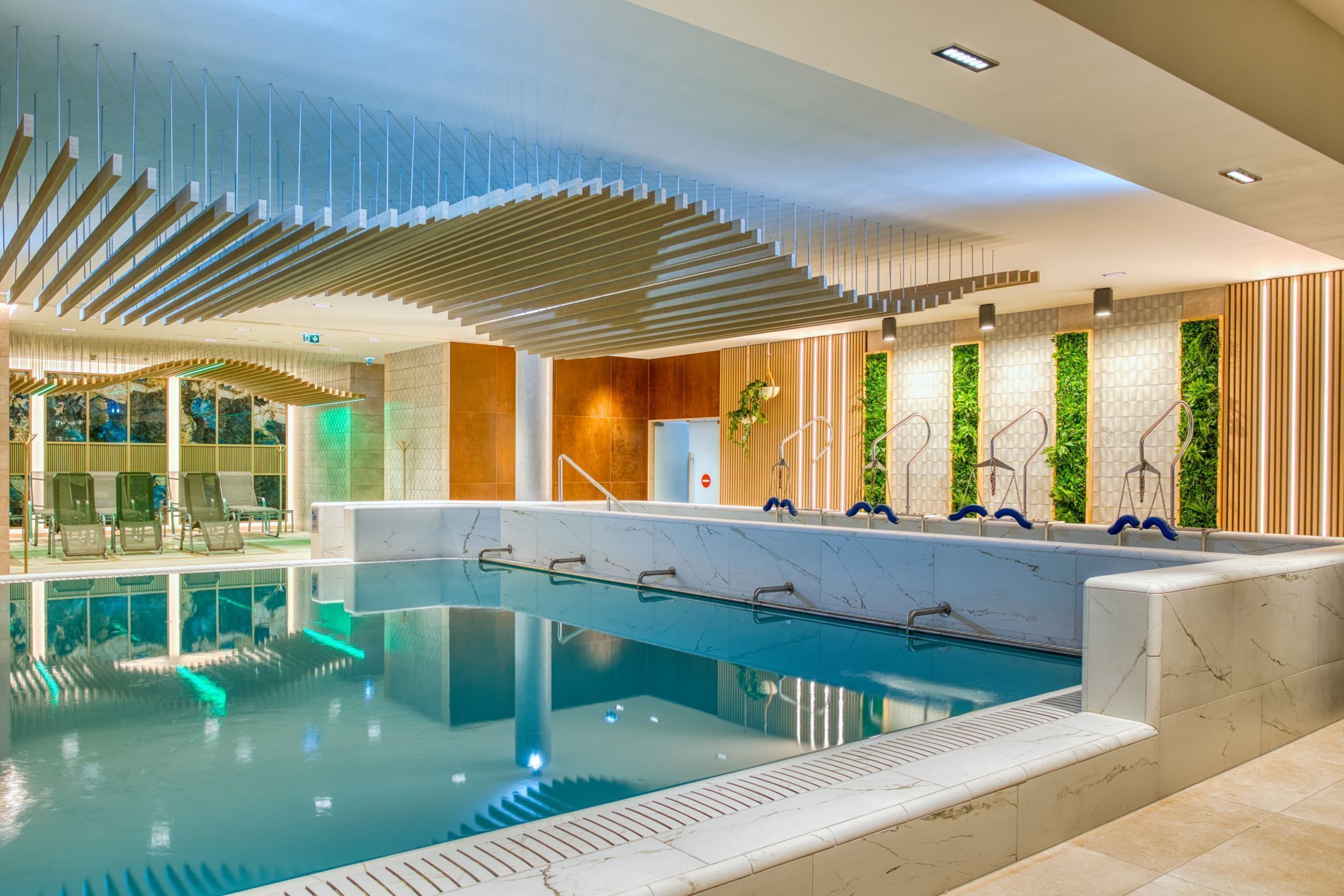Una piscina interior moderna con barrera de cristal, listones de madera en el techo y una zona de estar con sillas.