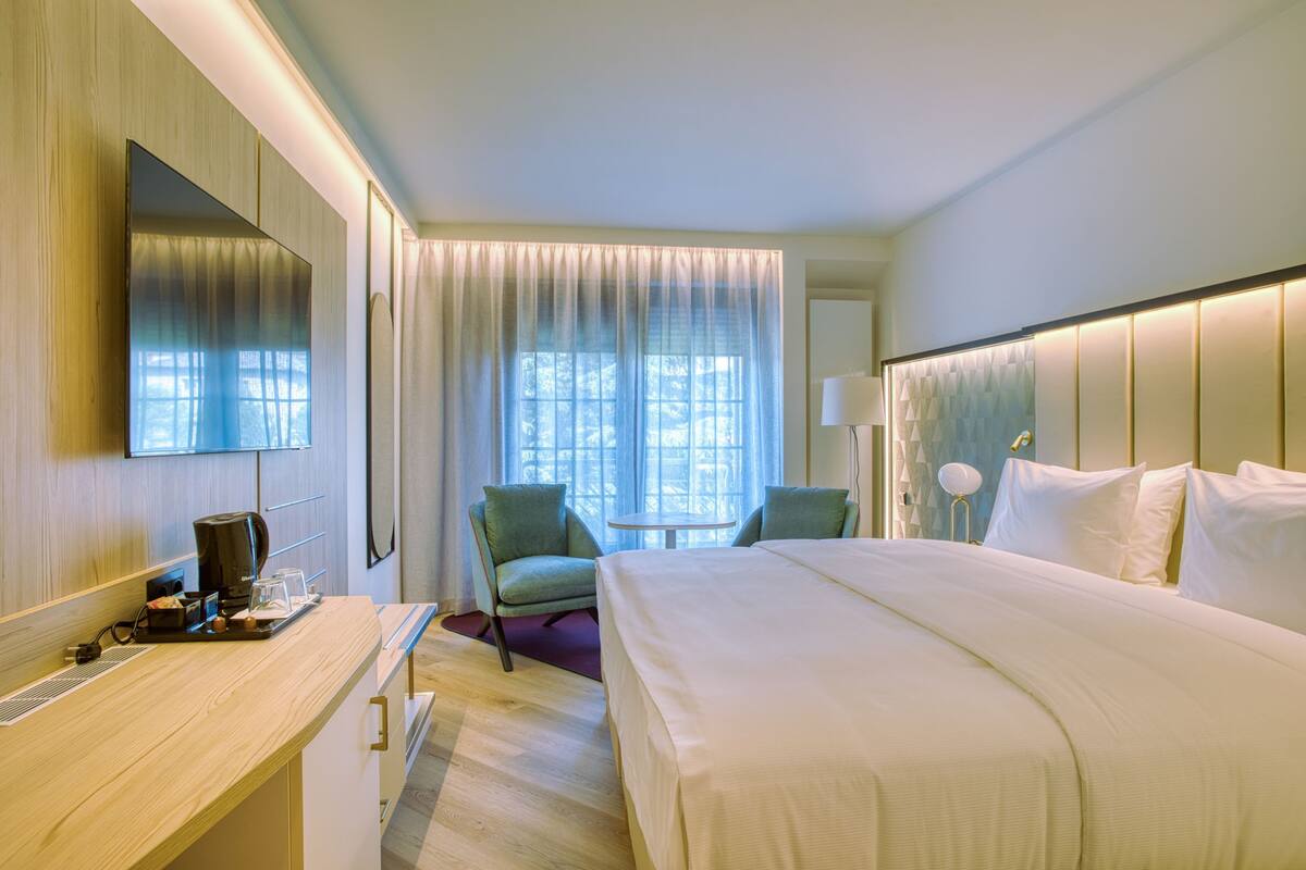 A hotel room with a large bed, a desk with a coffee maker, a chair, a round table, and a window with curtains.
