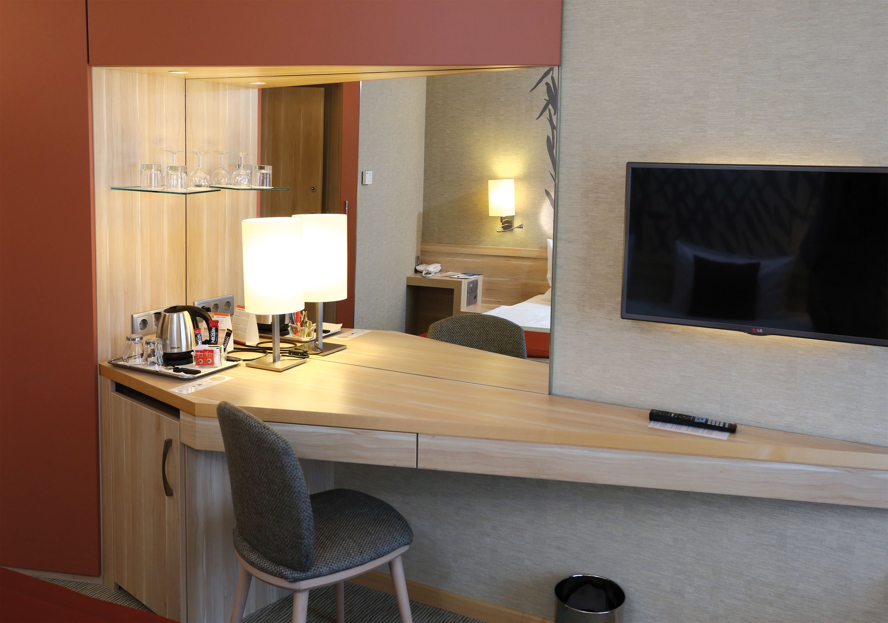 A hotel room with a wooden desk, a chair, a mirror, a TV, and a bedside table with a lamp.