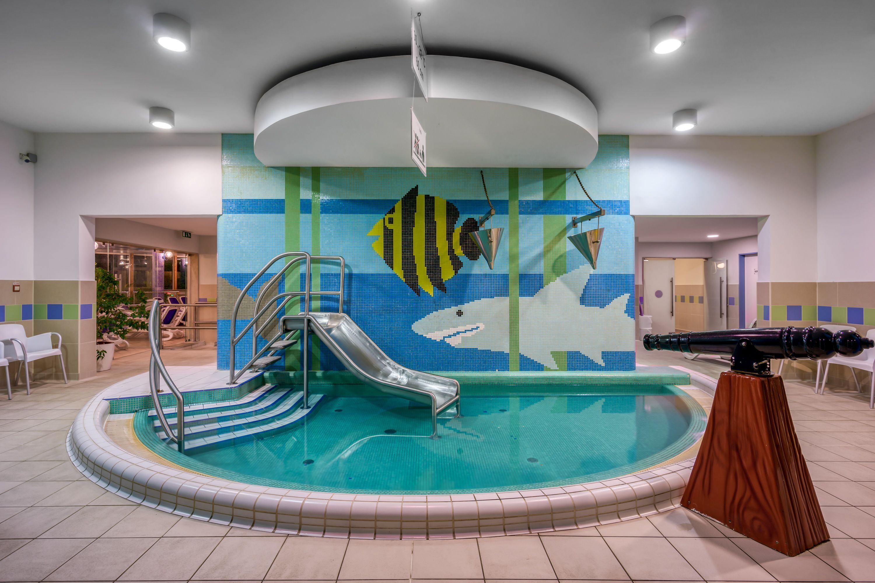 En inomhuspool med en vattenrutschkana, ett teleskop och en vägg dekorerad med en färgstark mosaik.
