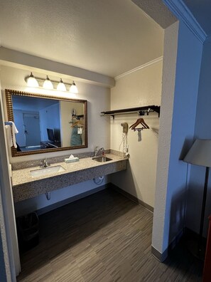 A bathroom with a double sink, a mirror with lights, a towel rack, and a hairdryer. - Studio 6 Fort Worth, TX – Downtown East (Fort Worth)