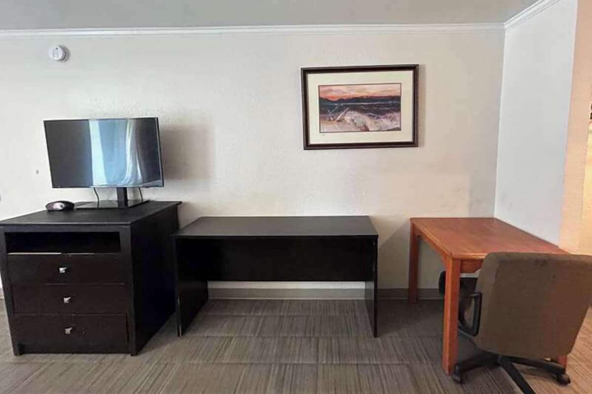 A small, sparsely furnished room with a desk, a chair, a television, and a framed picture on the wall.