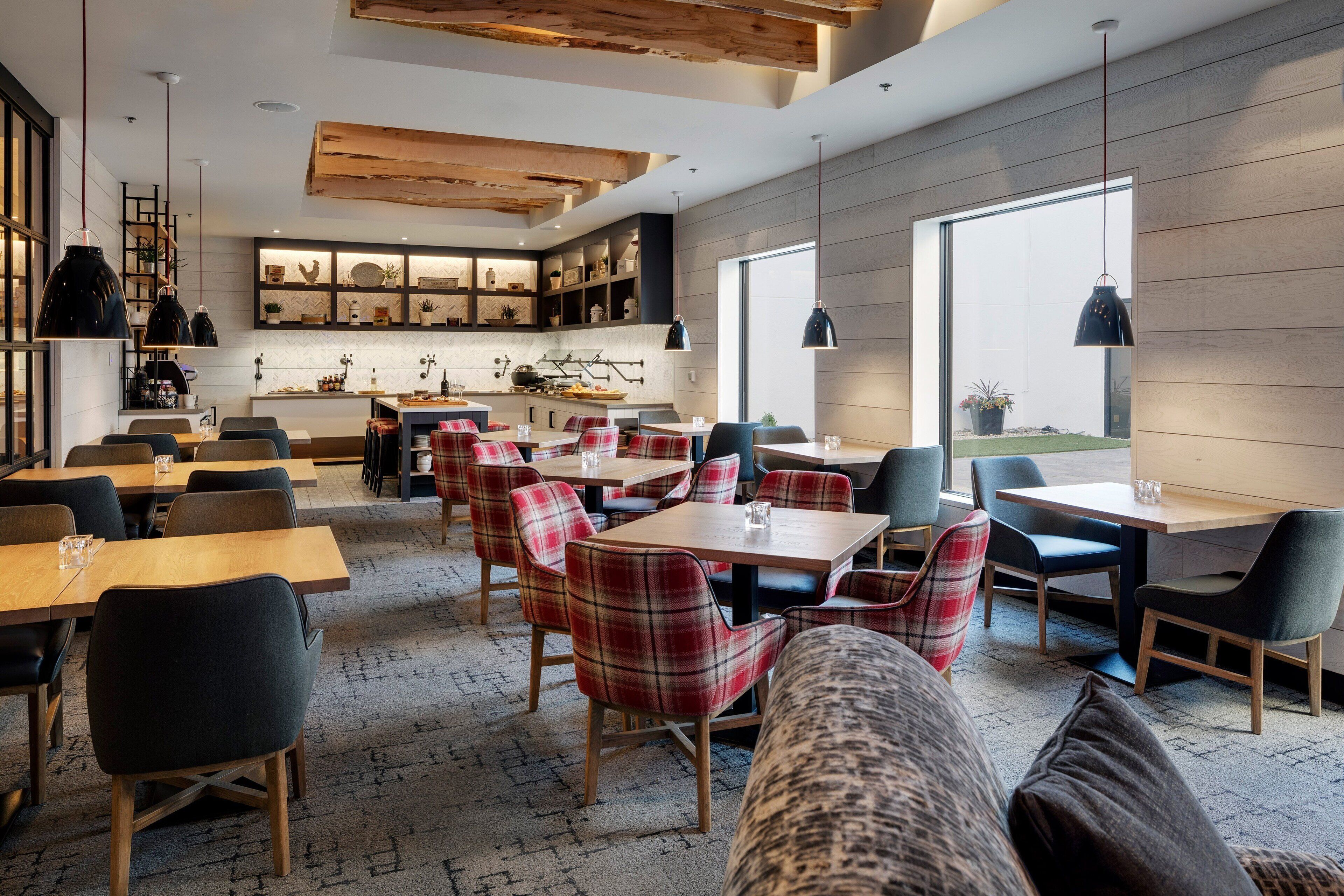 Een moderne eetruimte met houten tafels en geruite stoelen, een keuken op de achtergrond en grote ramen.
