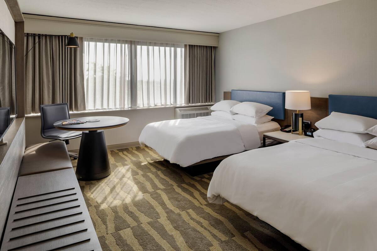 A hotel room with two beds, a desk, a chair, and a large window with curtains.