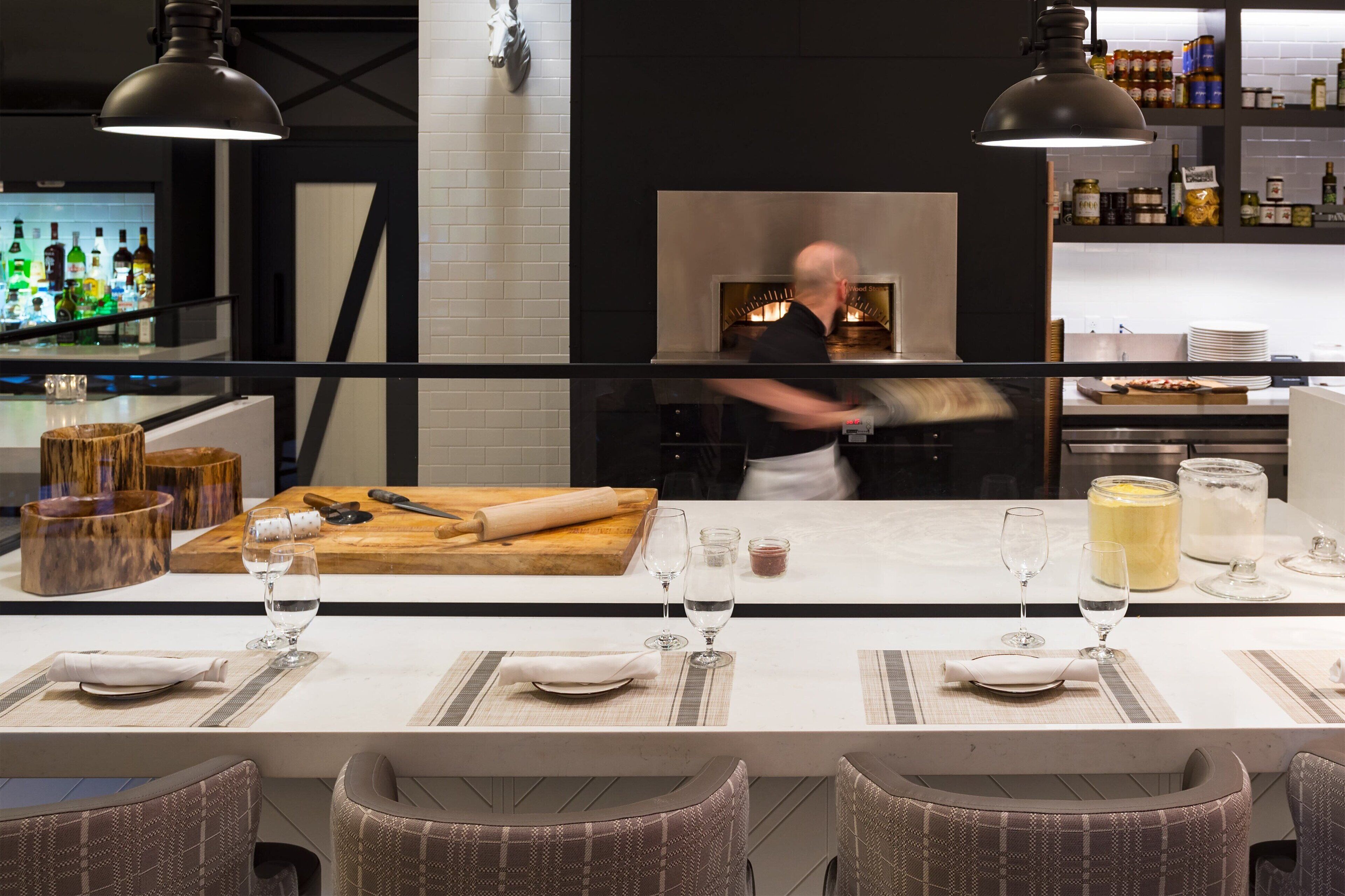 A modern kitchen with a chef preparing food, a bar area with bottles, and a dining setup with tables and chairs.