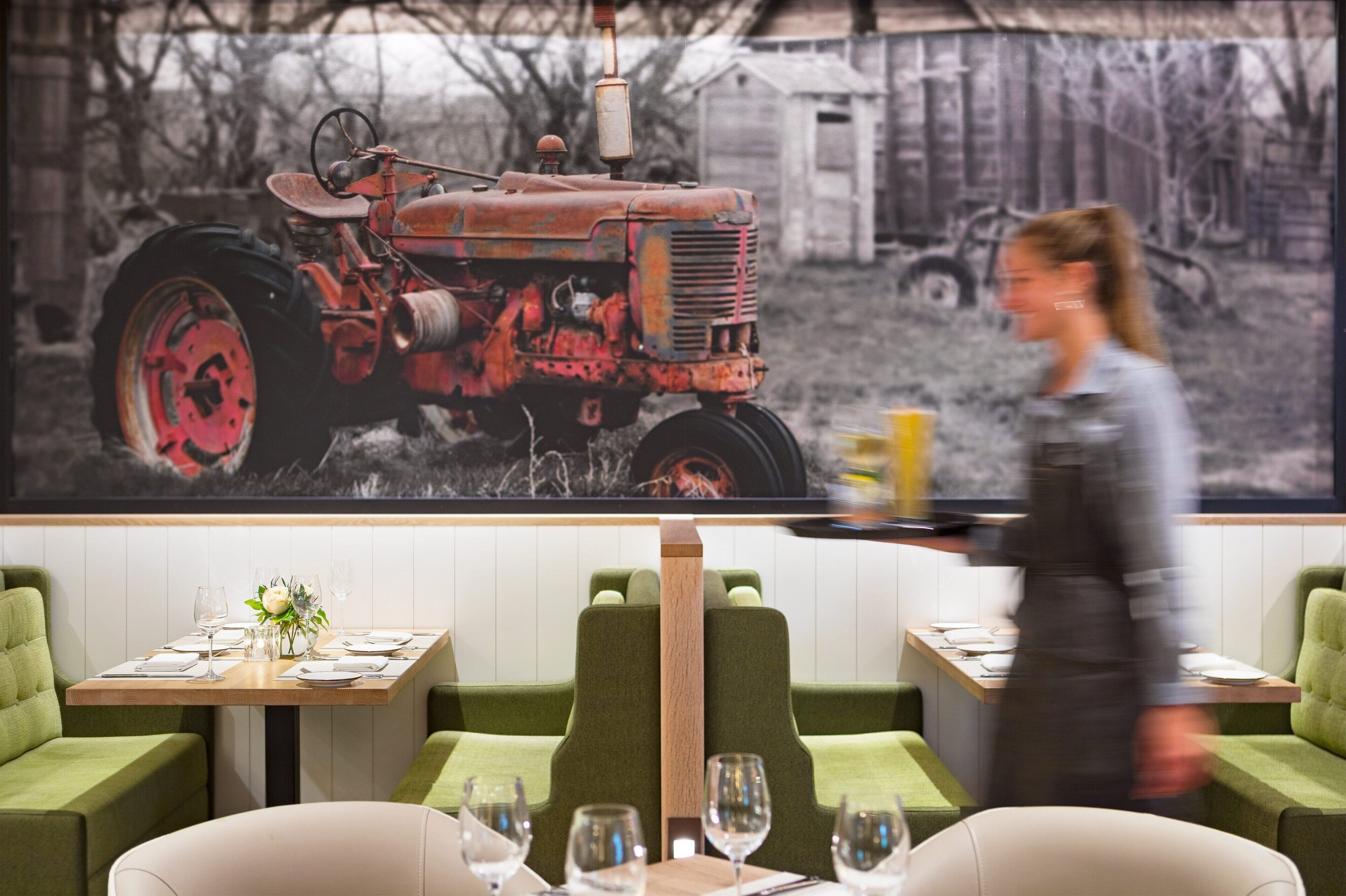 A restaurant with green upholstered booths, a table set with wine glasses and plates, and a large painting of a tractor in the background.