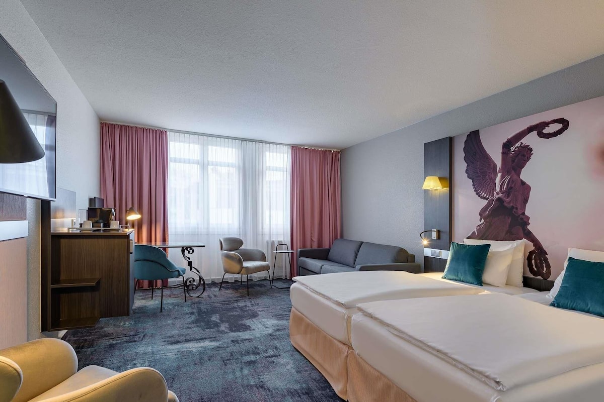 A hotel room with a large bed, a desk, two chairs, a sofa, and a large window with curtains.