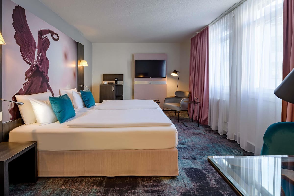 Een hotelkamer met een groot bed, een bureau, een stoel, een televisie en een muur met een groot kunstwerk.