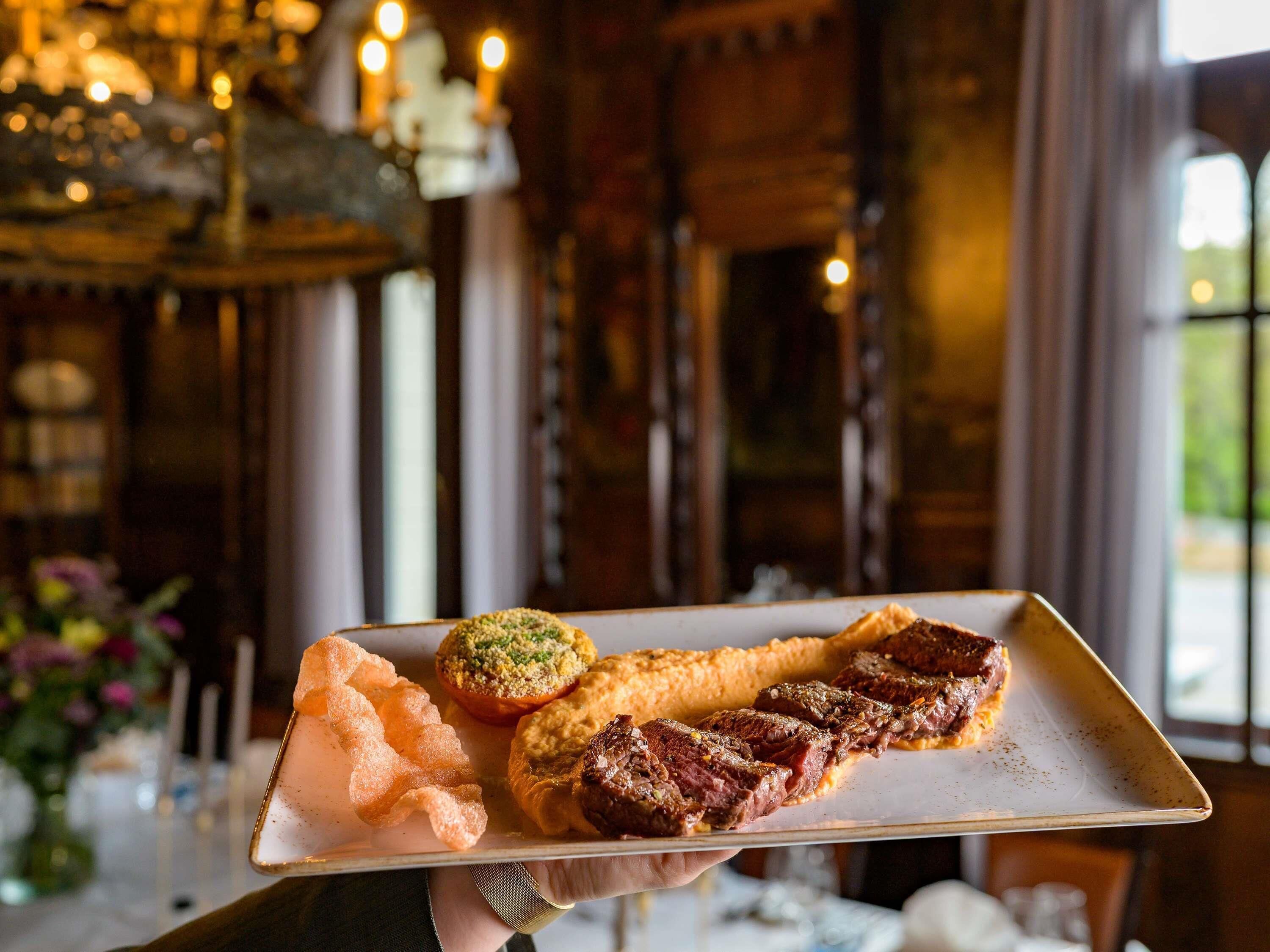 A person holding a tray with food items including a steak, a pastry, and a vegetable.