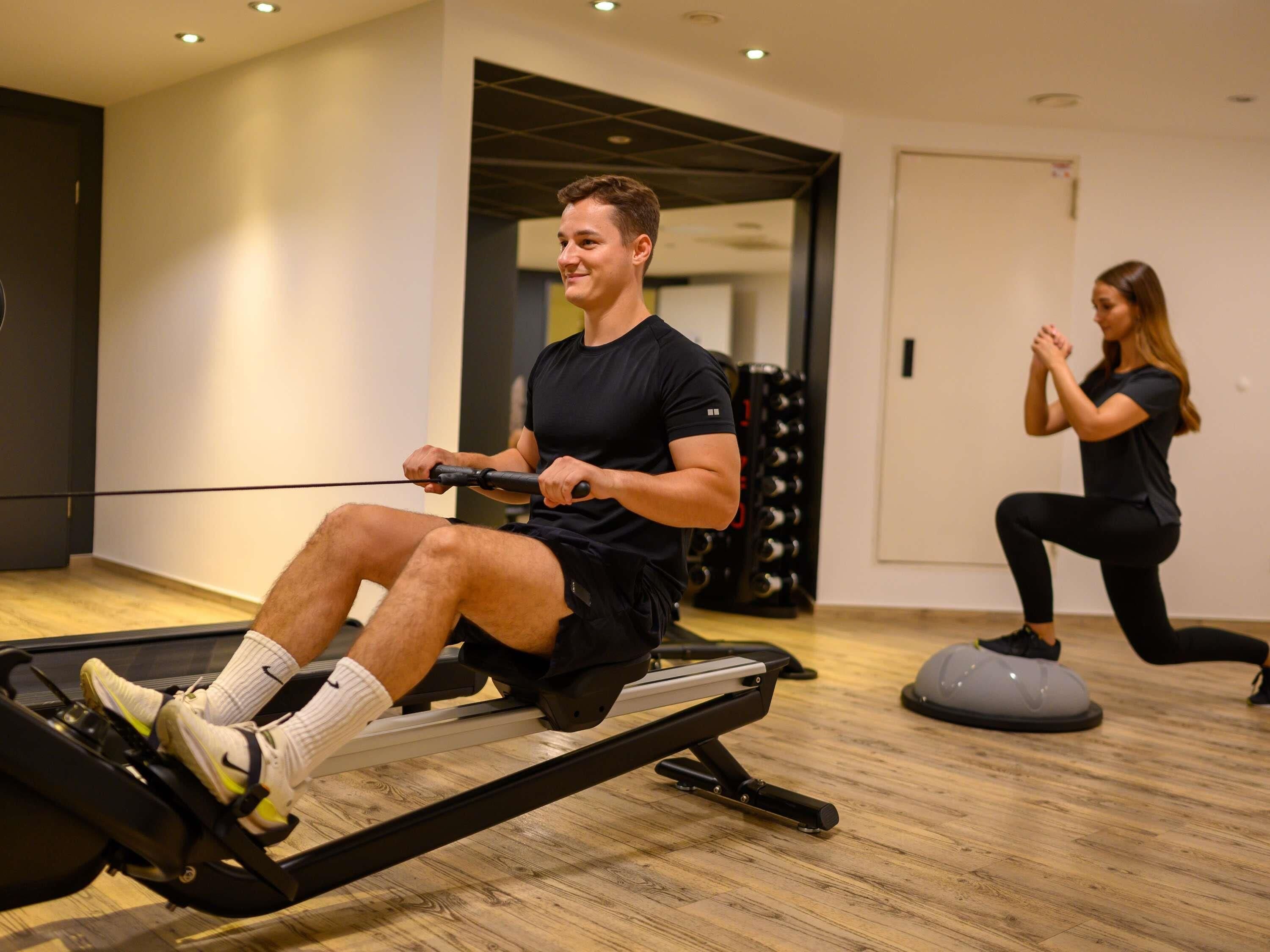 Un hombre usando una máquina de remo y una mujer haciendo sentadillas en un gimnasio con suelo de madera y diseño moderno.